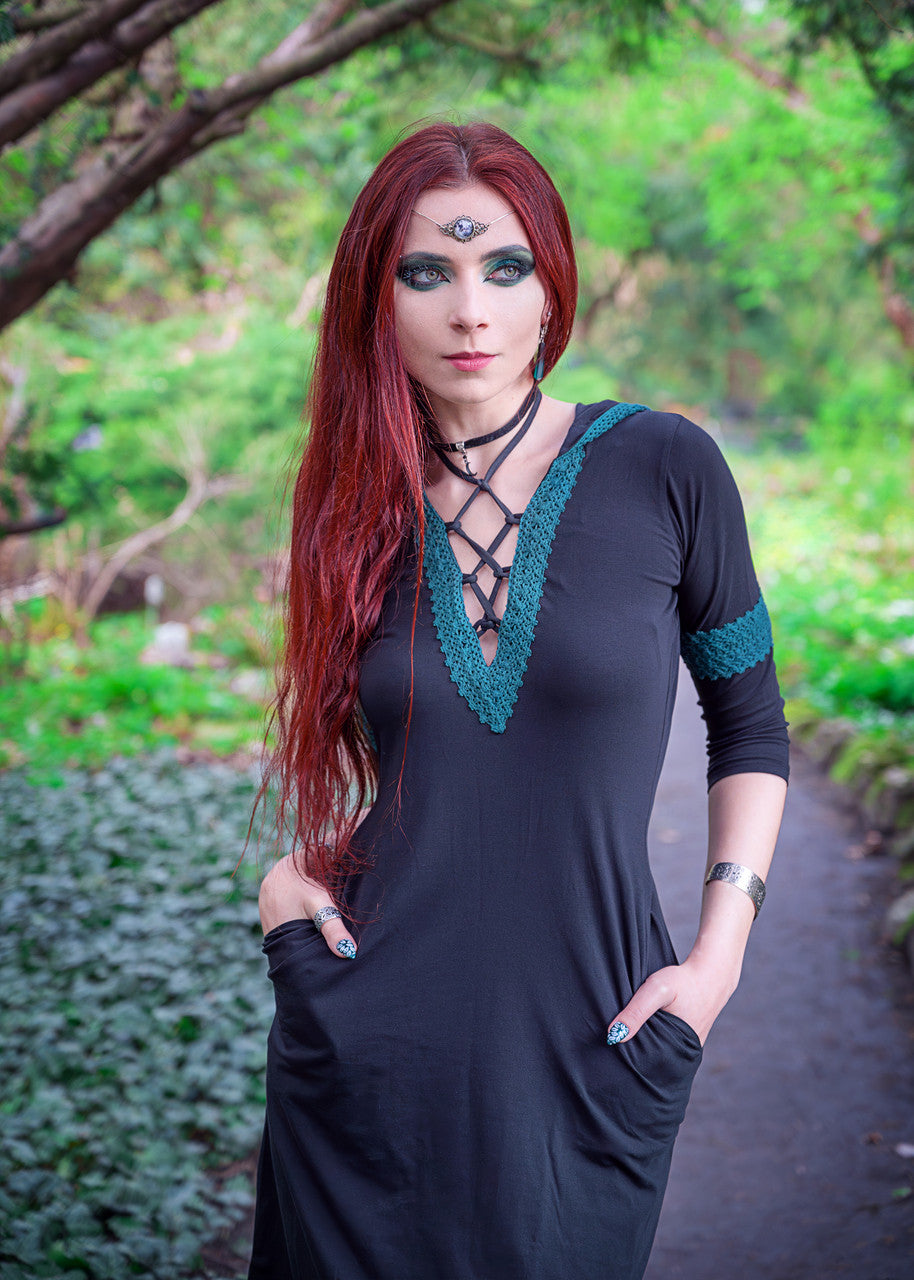 Woman posing in Dahlia Hooded Dress with both hands partially in inseam pockets, front view showing lace up chest , and forest green crochet lace surrounded by trees. @cyanide_red_model