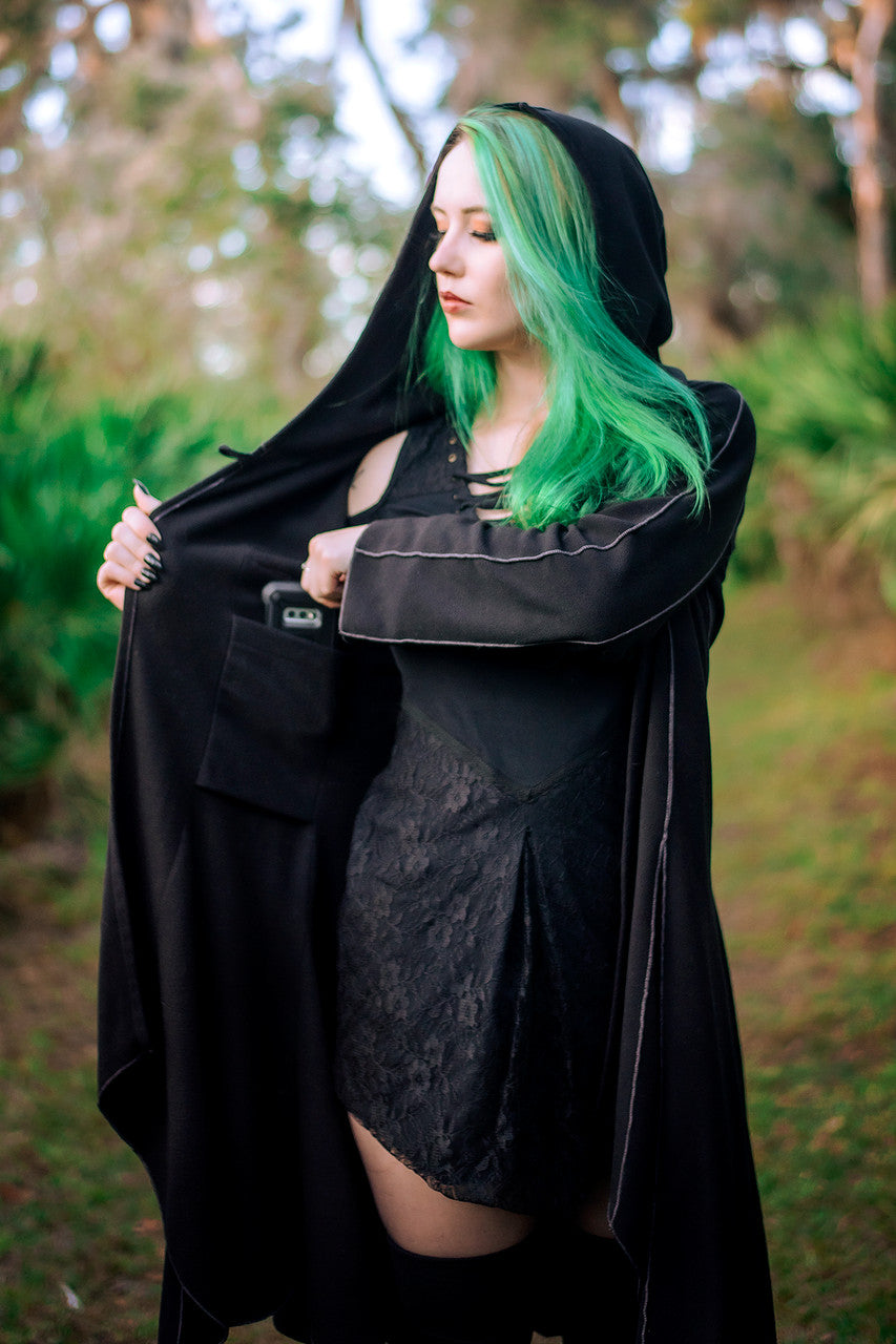 Conjurer's Coat in black hood up worn open and putting phone inside inner pocket, front angle.