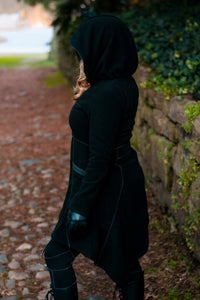 Model wearing black and green Necromancer's Coat, showing side view, on a cobblestone path, surrounded by greenery.