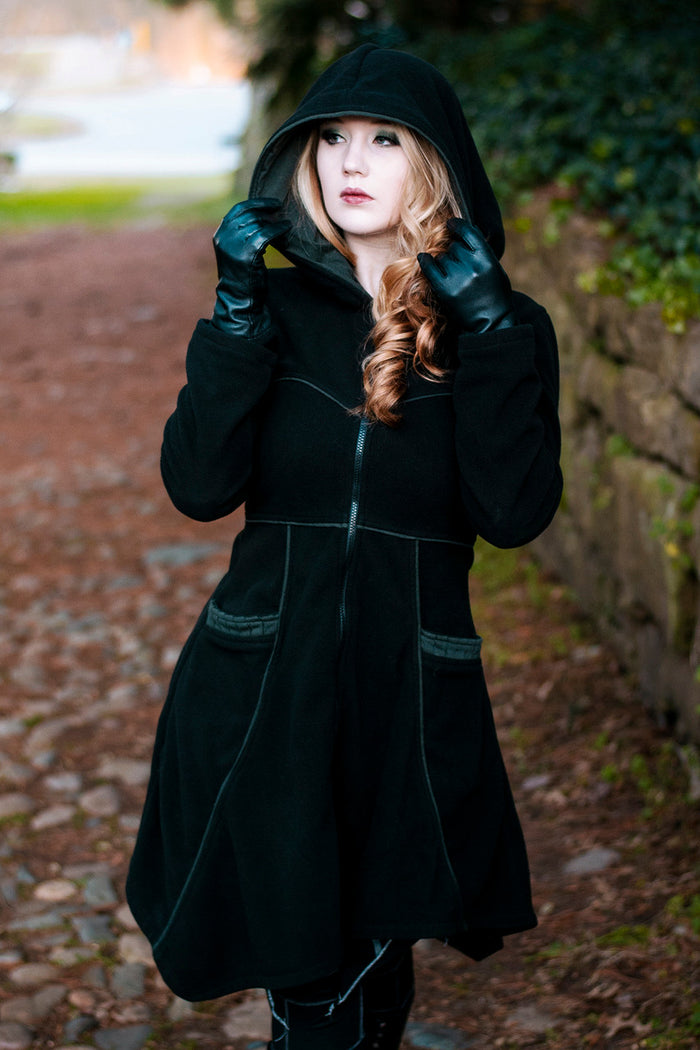 Model wearing black and green Necromancer's Coat with both hands holding the hood, on a cobblestone path surrounded by greenery.