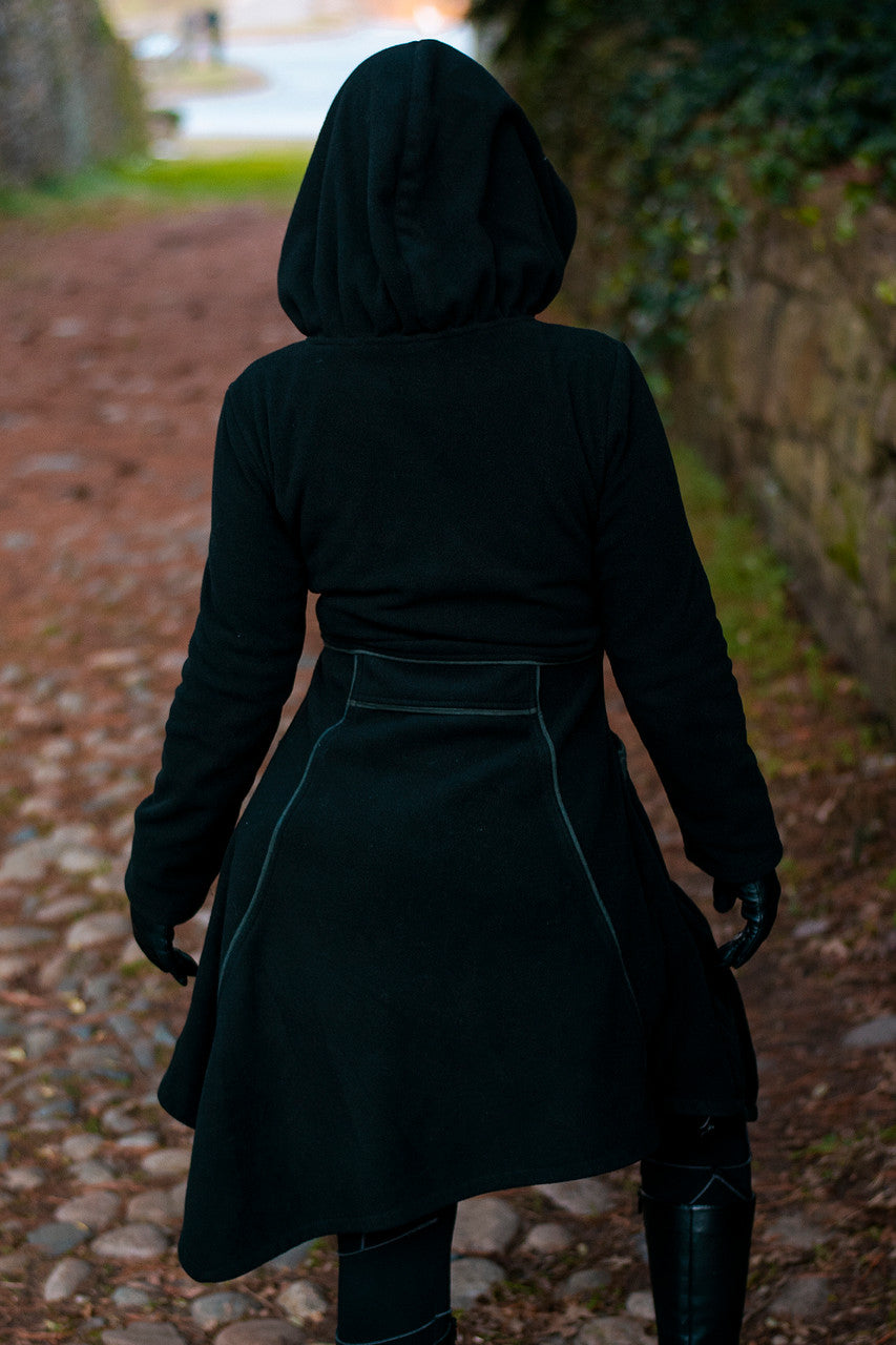 Model wearing black and green Necromancer's Coat, showing back view surrounded by greenery.