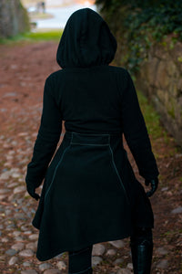 Model wearing black and green Necromancer's Coat, showing back view surrounded by greenery.