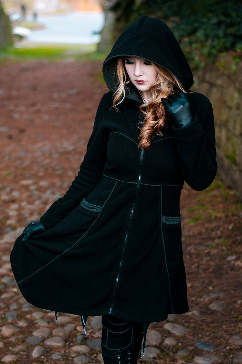Person wearing a black Necromancer's coat, walking down a cobblestone path surrounded by greenery, with a serene expression and one hand holding the hood.