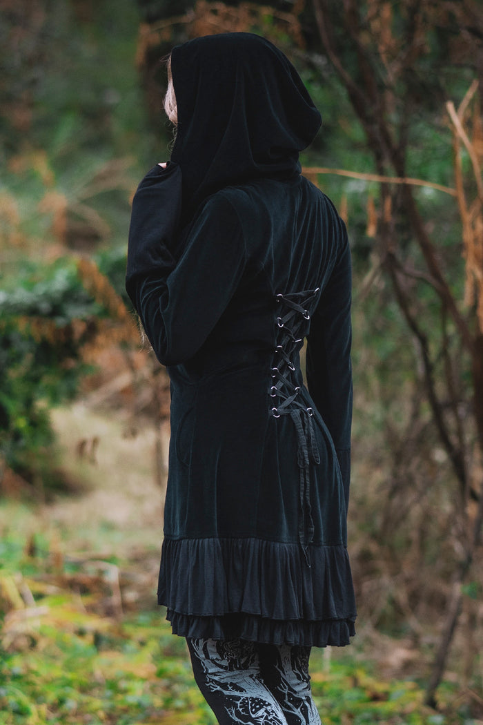 Model posing in woods, showing adjustable lace up back of the Occultist Coat and large wide hood.