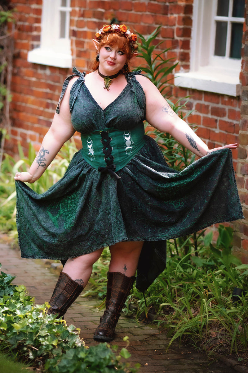 Woman in elf ears and flower crown posing in Morrigan Apron Dress in black and green  in front of brick building, holding out the ends of the dress. The dress is paired with forest green Selene Corset.
